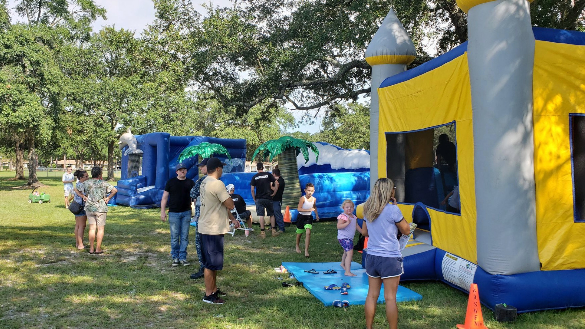 Image of three inflated bounce houses on green grass with people in and around them. Background is trees.