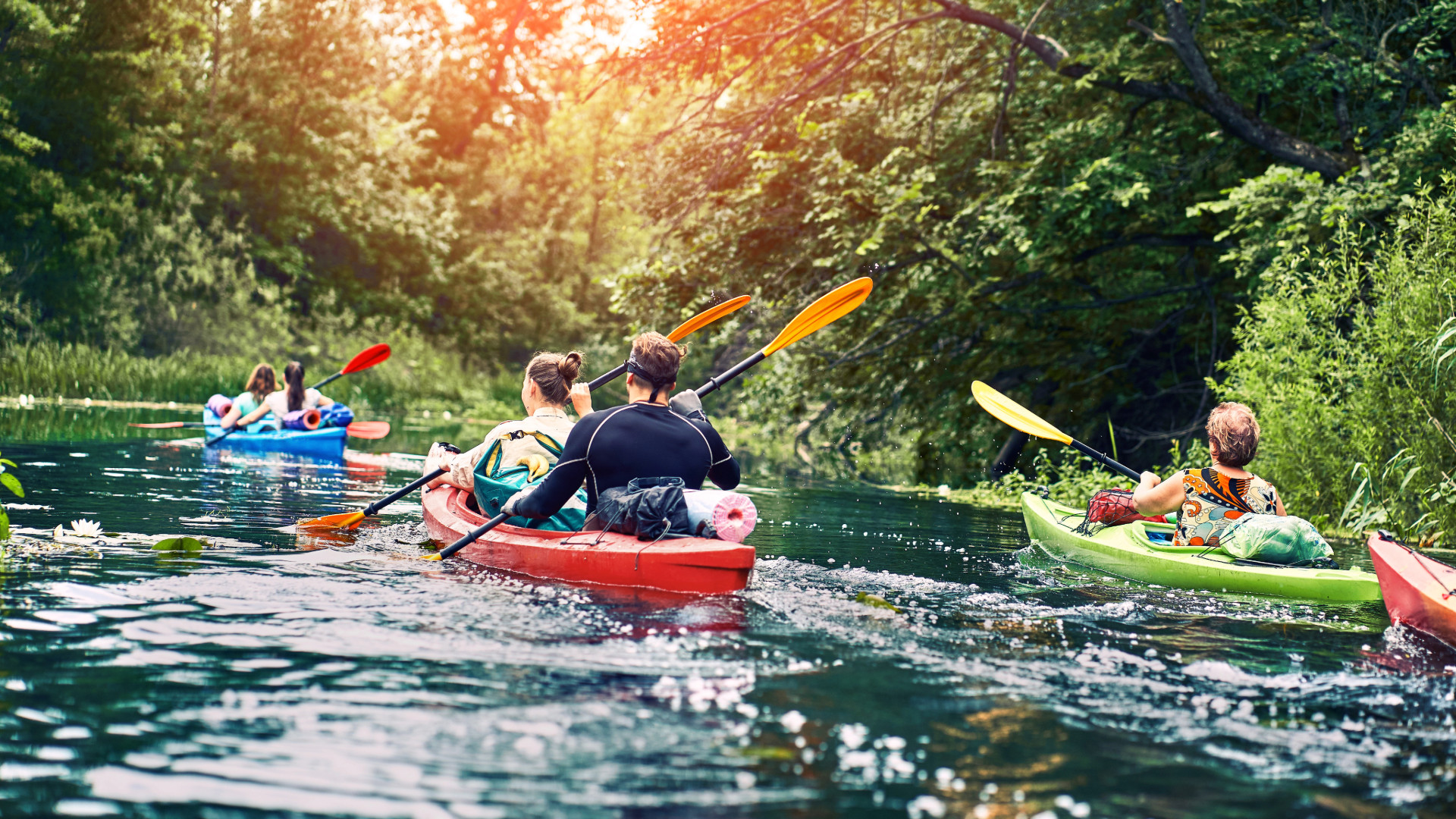 Image of five people spread between three kayaks on a river. Sun is shining through the trees above.