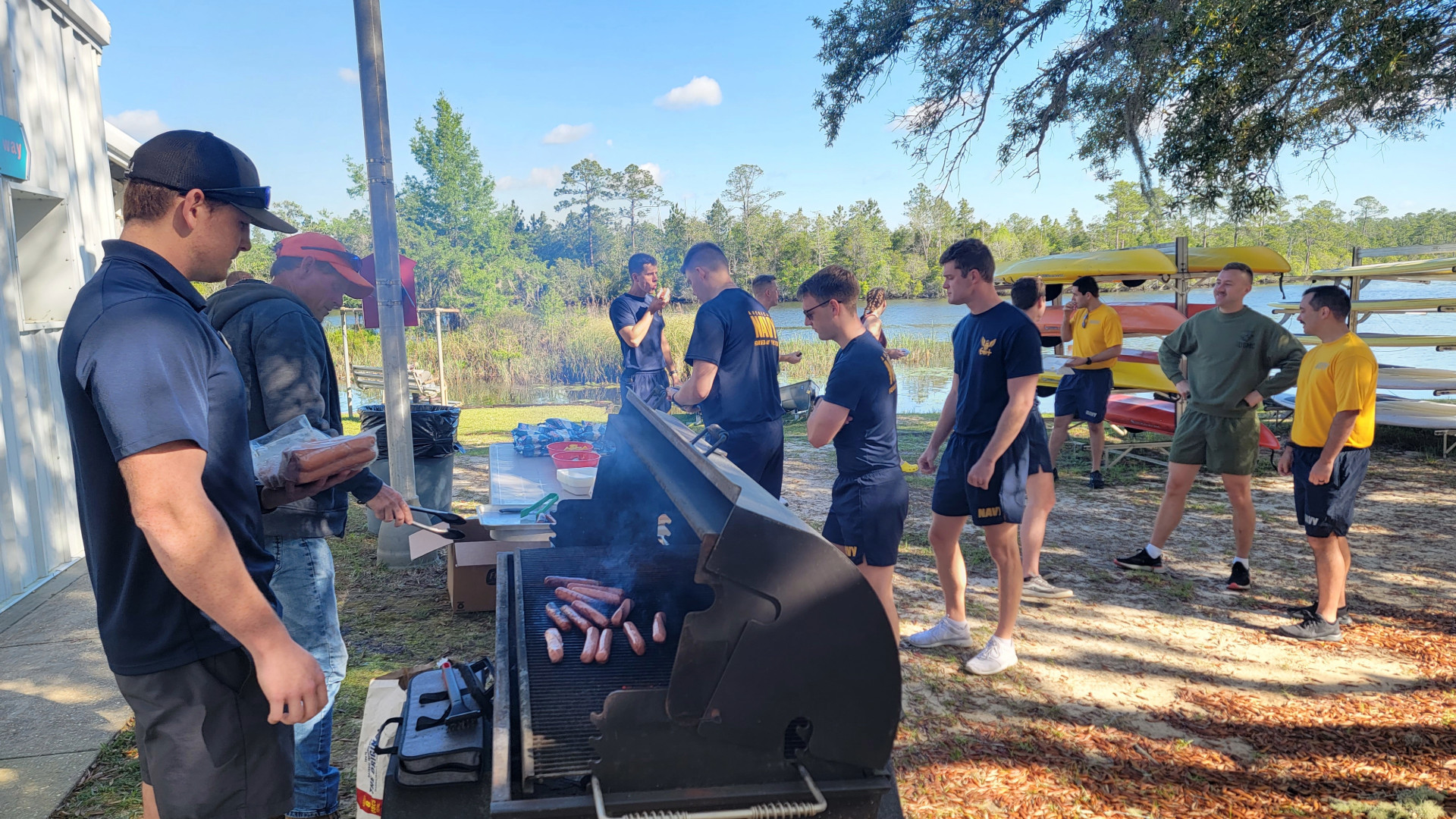 Photo of two people grilling hot dogs by a river. A crowd of people in blue t-shirts and shorts stand around. Kayaks are stacked in the background.