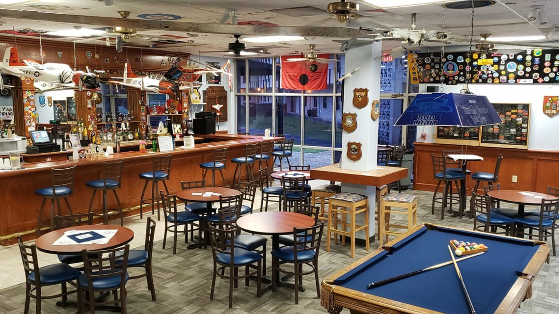 A cozy bar featuring round wooden tables, blue cushioned chairs, a pool table, and aviation-themed decor. The walls display patches and flags.